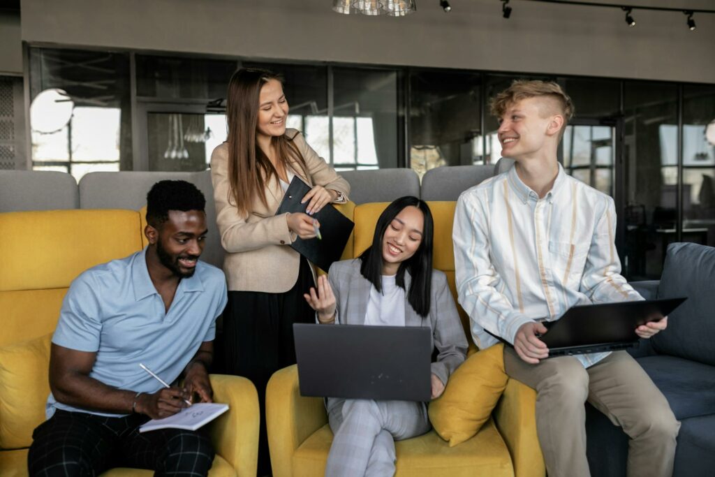 group of four co-workers, two women and two men, sitting in a yellow coach while working. Each of them is holding a computer/tablet.