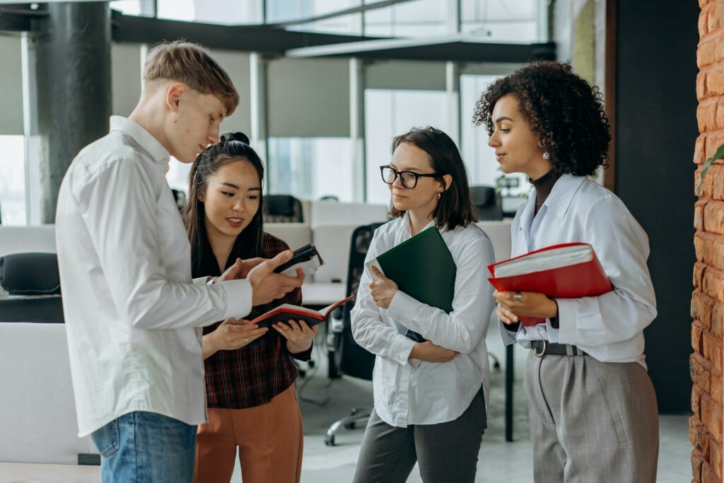 Group of 4 coworkers standing in an office hallway, discussing and comparing notes.