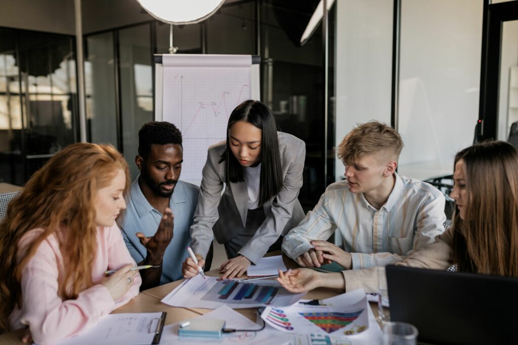 A diverse group of five young professionals collaborating around a conference table in a modern office, looking closely at printed charts and graphs.